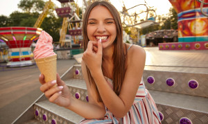Cheerful young beautiful female with long brown hair sitting over park of attractions with ice cream in hand, looking to camera with broad smile and keeping thumb on her underlip